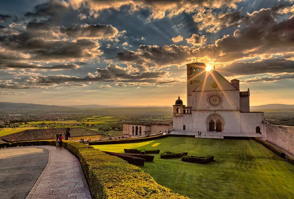 Basilica of St.Francis in Assisi, Italy, sunset over the cathedral, green grassy patch, dark clouds in sunslight