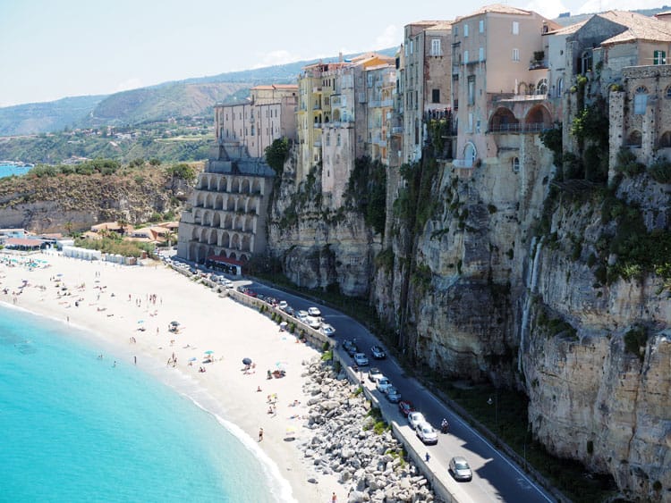 Italy Tropea, very high rocky cliff with buildings on the tops, road below with cars parked next to the beach, beach with people on it, light blue water