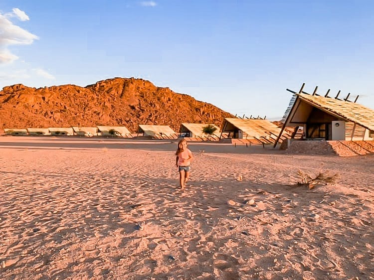 Camping in a desert with Kids - Namibia Desert Quiver Camp, young girl running in the desert, villas and a rocky hill in background