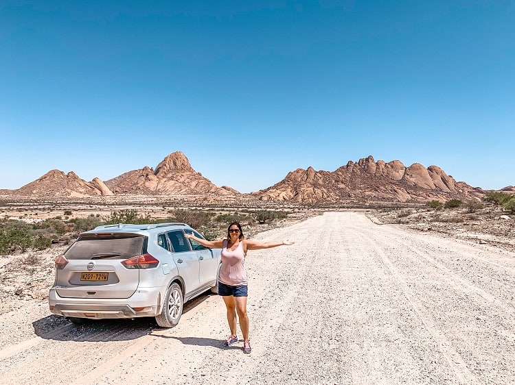 Driving in Spitzkoppe, Namibia, woman posing arms up next to the car on the dirt road, rocky mountains in background